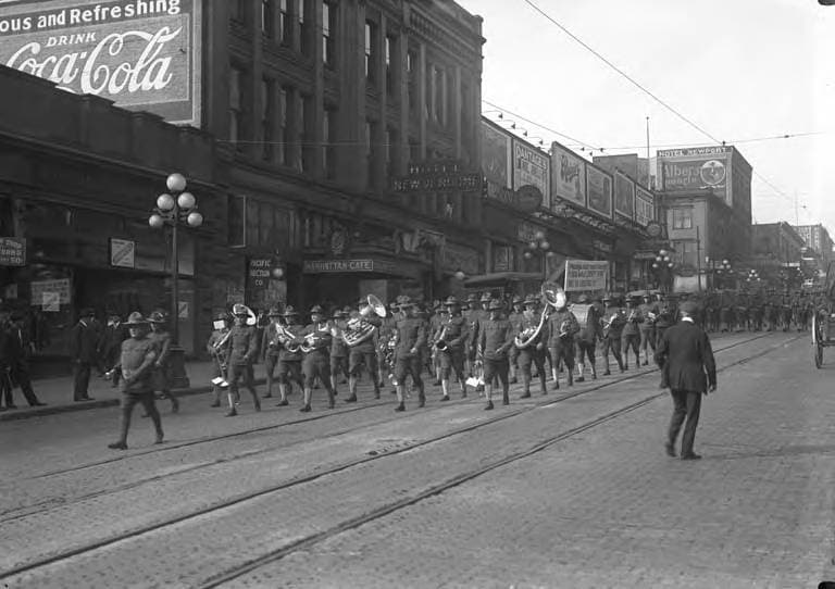 Black-and-white archival photograph of a military band marching on 1st Avenue, Seattle, around 1917 to 1920, with the Hotel New Vendome sign and the Manhattan Cafe storefront behind them