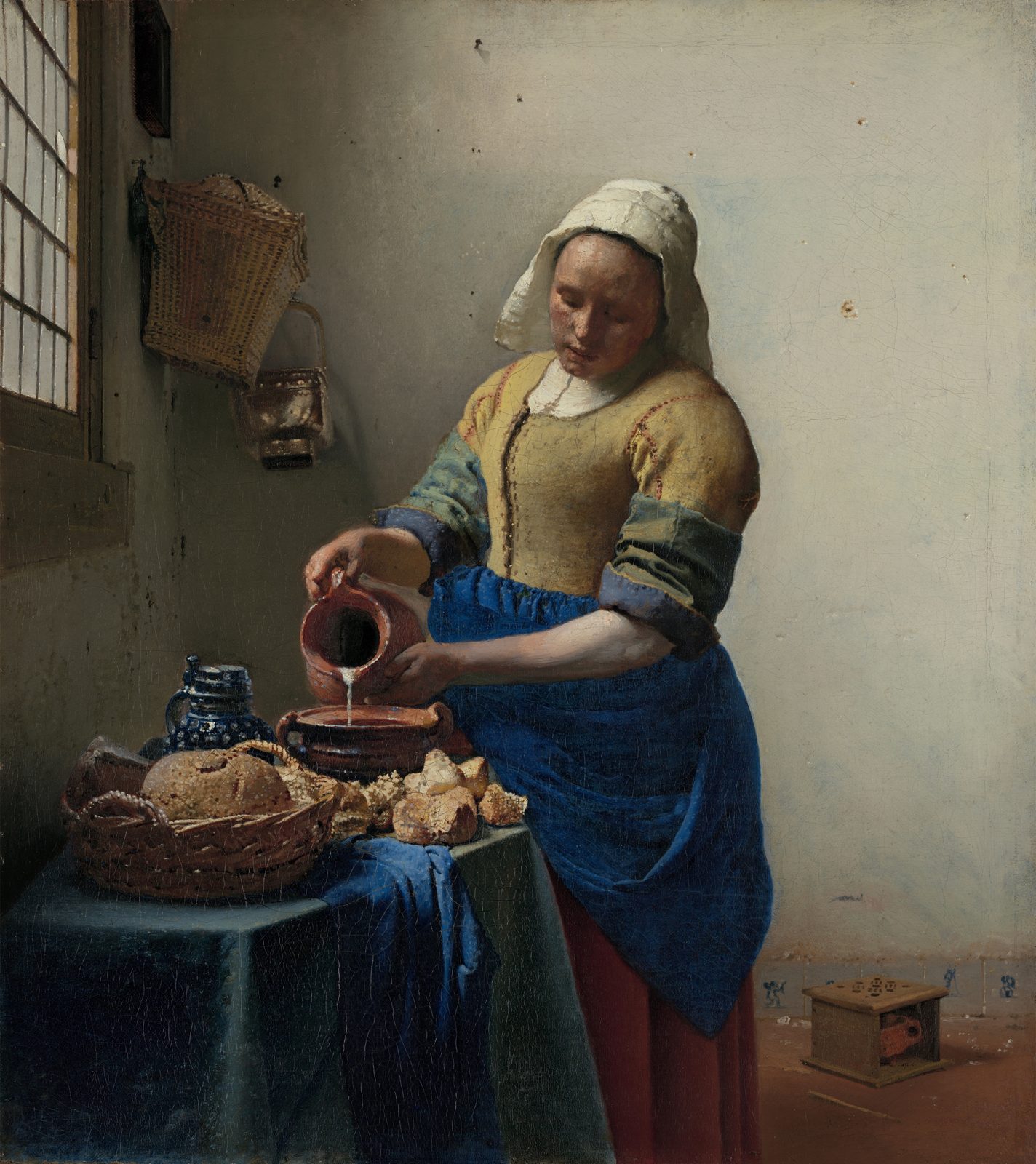 A kitchen maid in a yellow bodice pours milk from a jug into a bowl by a window, bread on the table before her