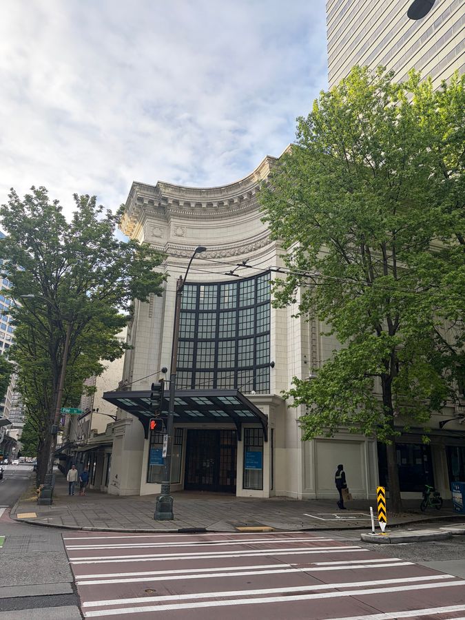The whole building from the corner of Fifth Avenue and Pike Street: the curved cornice, the tall grid window and the glass canopy. May 25, 2026.