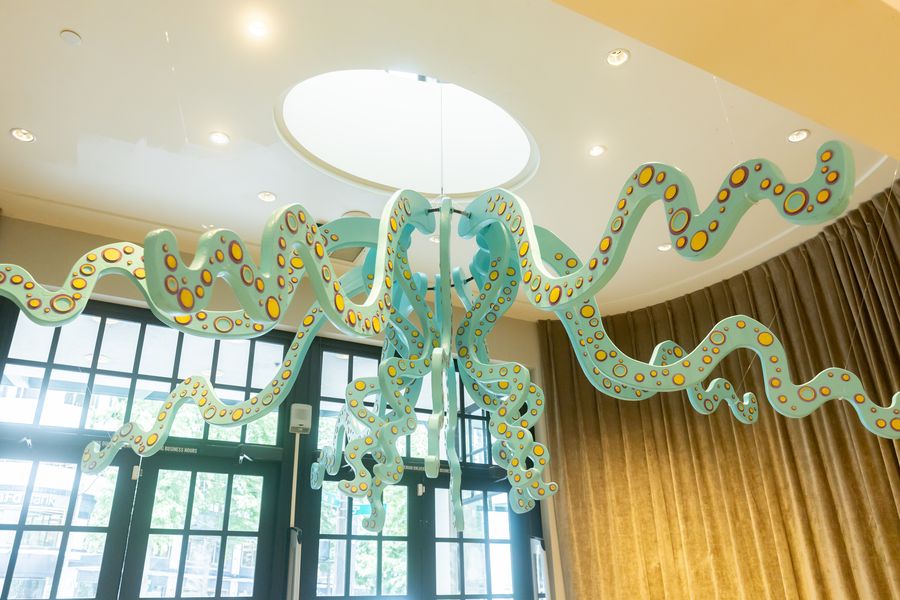 A sea-green tentacled sculpture suspended under the round skylight, with gold drapes and tall windows behind. July 7, 2026.
