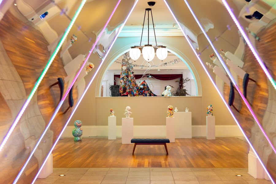 The long gallery hall seen through converging neon light lines: sculpture on plinths, globe chandeliers, an arched portal. July 7, 2026.