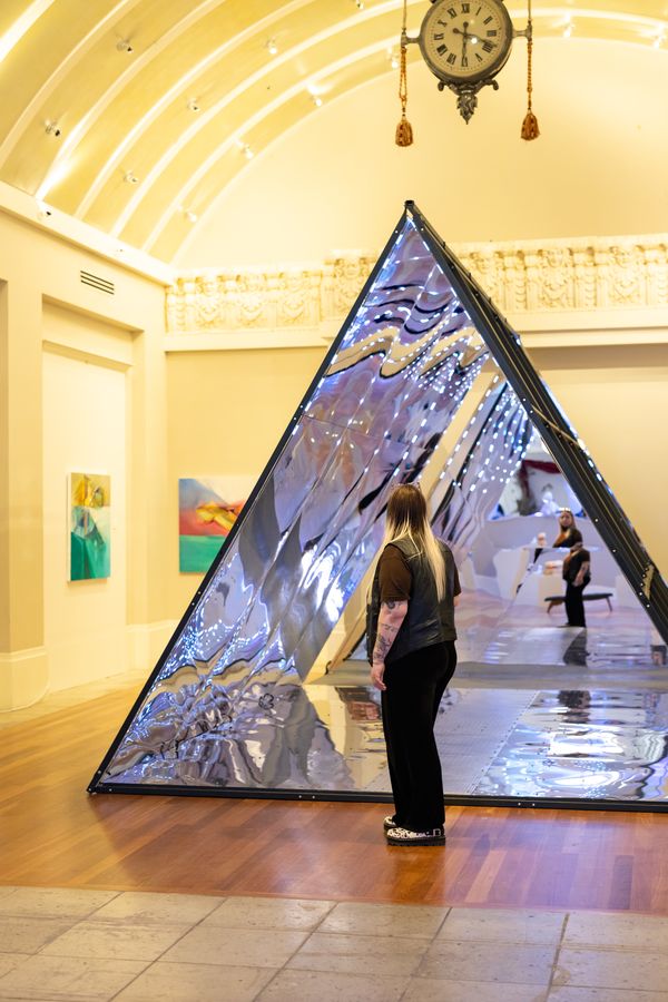 A visitor at the mouth of the mirror pyramid, with the clock and vaulted ceiling above. July 8, 2026.