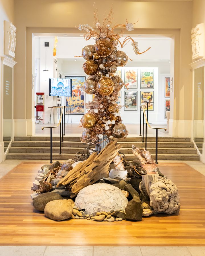 A tall amber found-object tower on a river-stone base in the main hall. July 8, 2026.